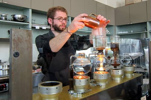  A barista adding coffee grounds to the top chamber of the siphon device