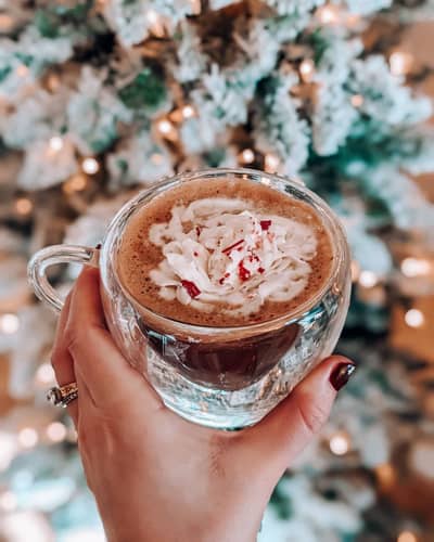 Closeup of woman holding a mug of Peppermint Mocha in front of a lit Christmas tree