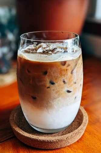 Iced version of a cappuccino topped with milk foam in a clear glass on a cork saucer