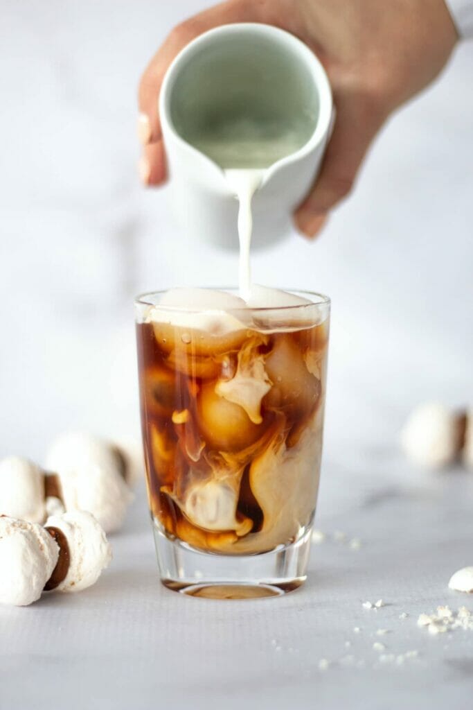 A hand pouring milk in a clear drinking glass with ice cubes