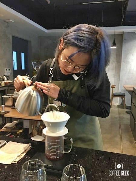 Aeropress vs V60: Woman pouring liquid from a kettle into a glass coffee mug