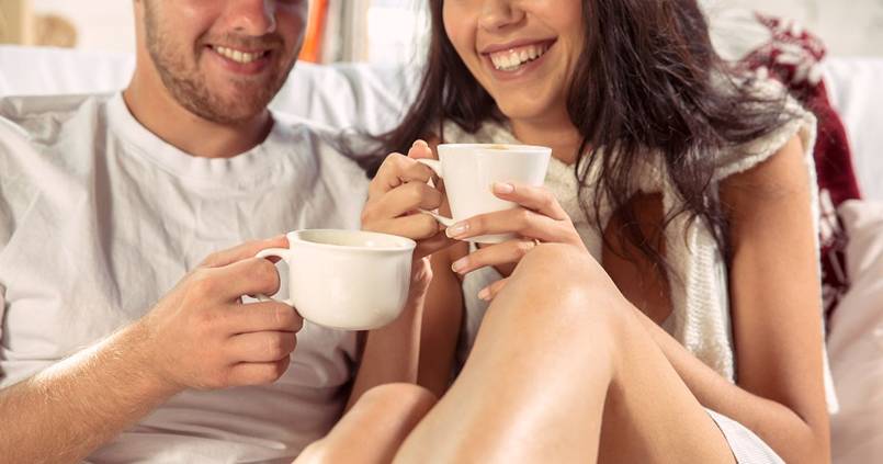 A man and woman are sitting on a bed and drinking coffee.