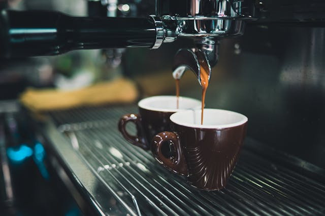 Two brown ceramic cups are placed under a coffee machine, with espresso being poured into both cups simultaneously.