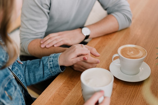 Two people sit at a wooden table holding hands, with two cups of coffee in front of them. One person is wearing a denim jacket, the other a light sweater and a watch.