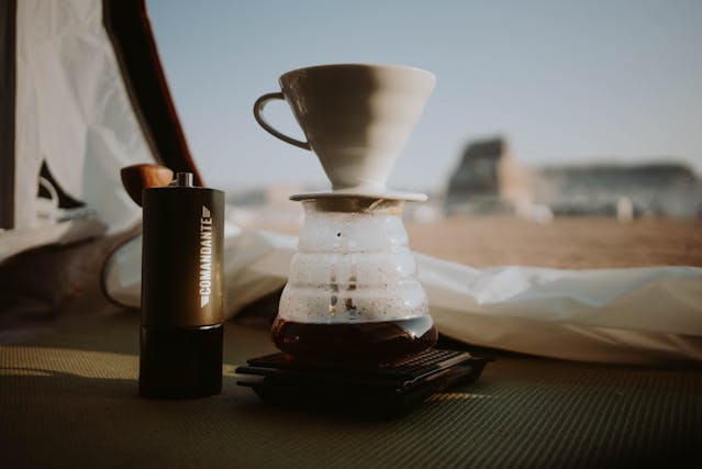 A manual coffee grinder and a pour-over coffee setup sit on the ground inside a tent, with blurred outdoor scenery visible in the background.